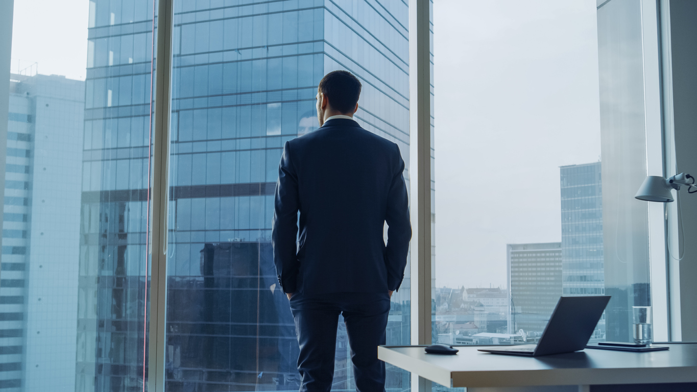 Back View of the Thoughtful Businessman wearing a Suit Standing in His Office, Hands in Pockets and Contemplating Next Big Business Deal, Looking out of the Window. Big City Business District Panoramic Window View.