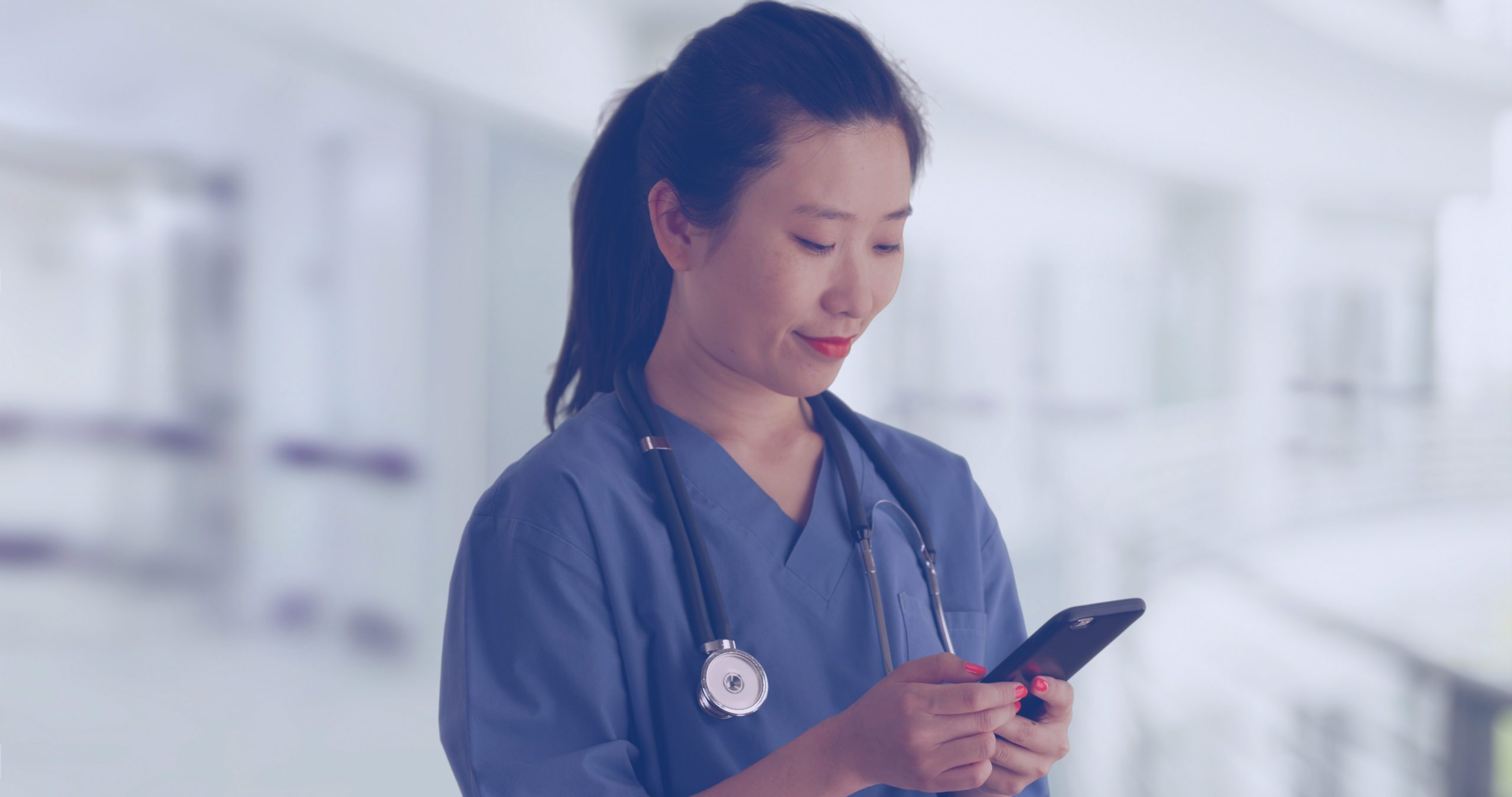 Happy young Asian doctor in scrubs texting with smartphone inside empty hospital